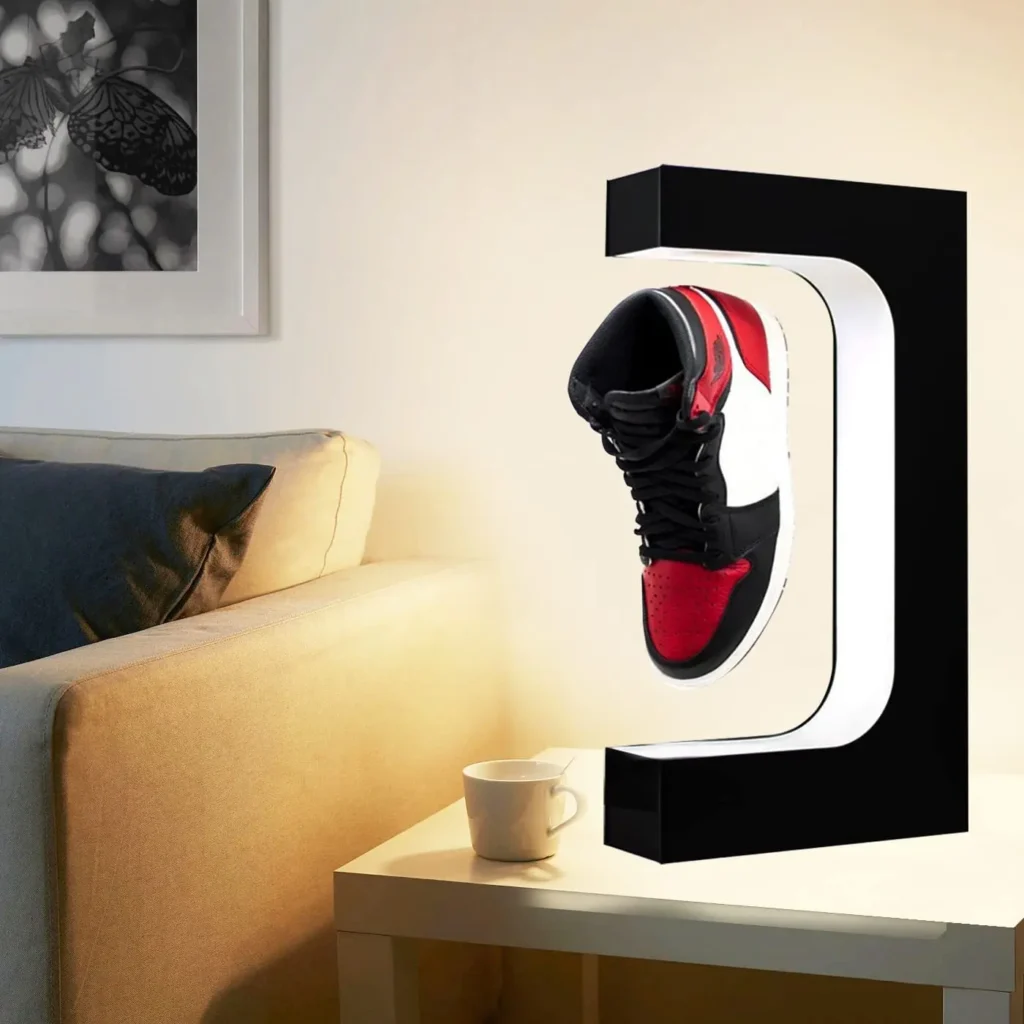 A single black, red, and white sneaker is displayed in a modern, illuminated frame beside a beige couch with pillows and a white coffee cup on a table.