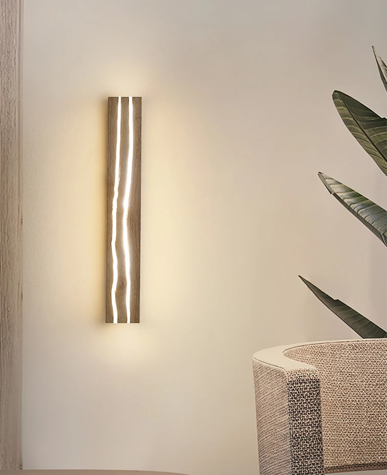 A vertical wall-mounted light fixture with a wavy design illuminates a neutral-colored room. A plant and part of a beige chair are visible.